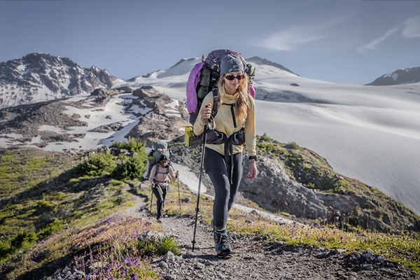Two women hikers with backpacks on a mountain trail with snow-capped mountains in the background.