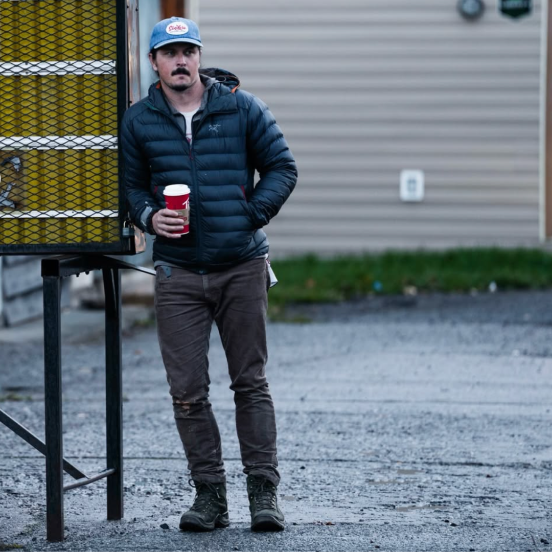 Man in a blue jacket and cap holding a red cup, standing on a gravel surface with a building in the background.