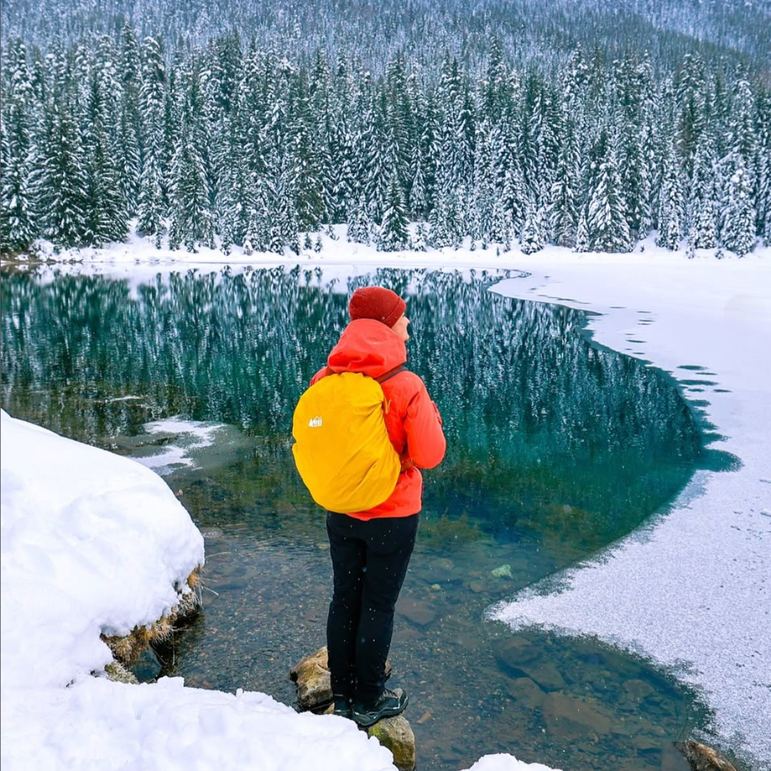 Person in red jacket and yellow backpack standing on a snow-covered landscape with a lake and trees.