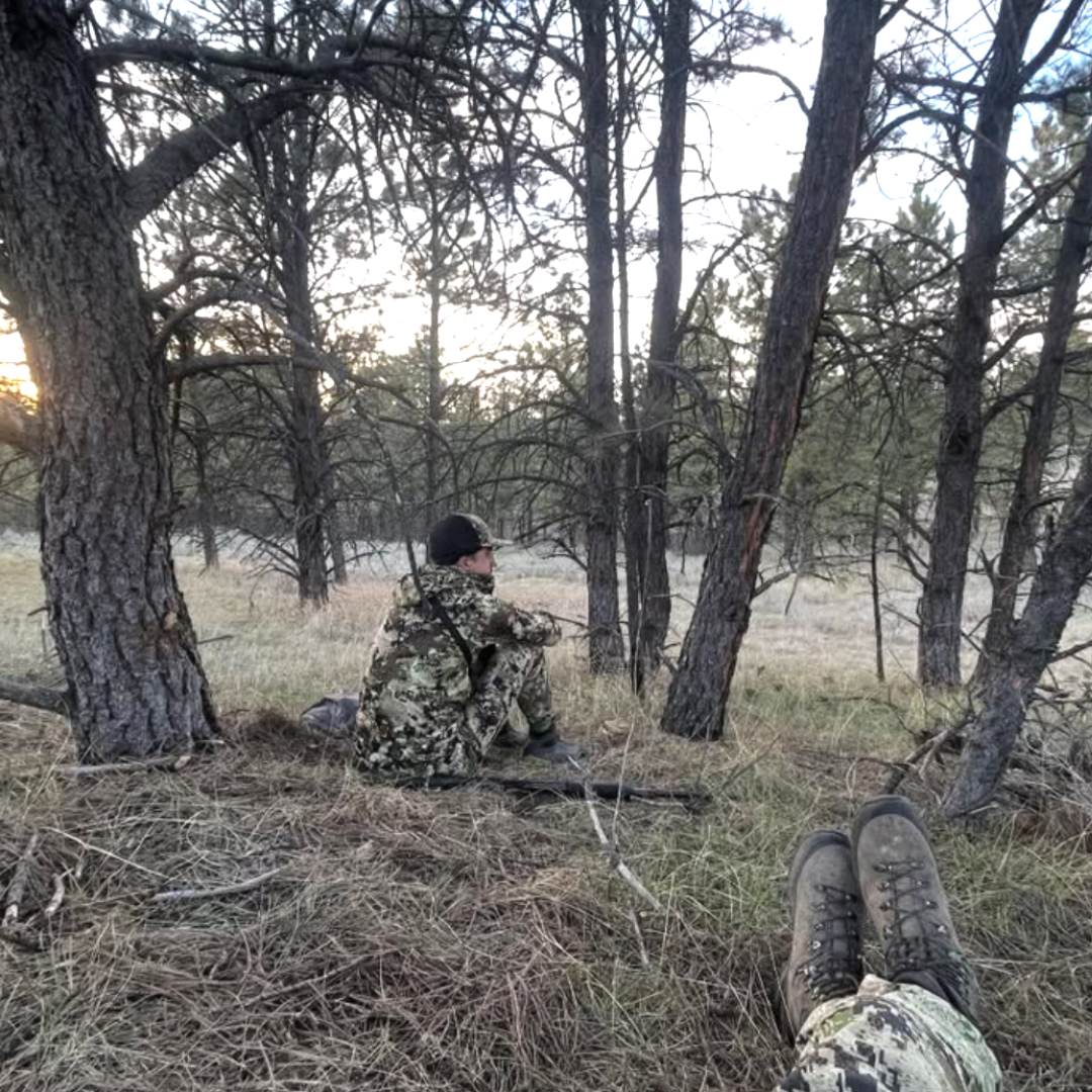 Person in camouflage sitting in a forest with a rifle, another person's boots visible in the foreground.