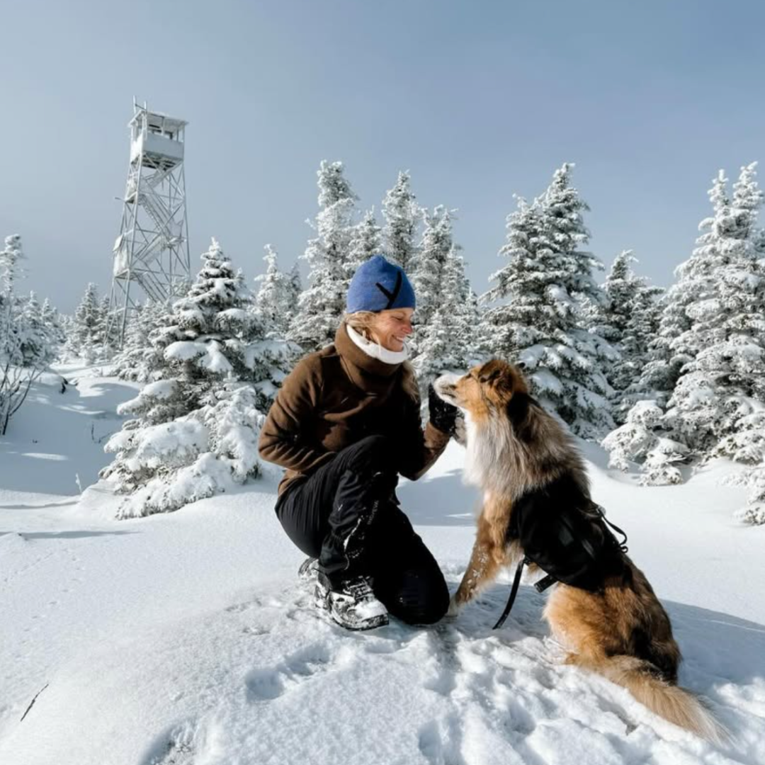 Person in winter clothing kneeling in the snow with a dog, surrounded by snow-covered trees and a tower.