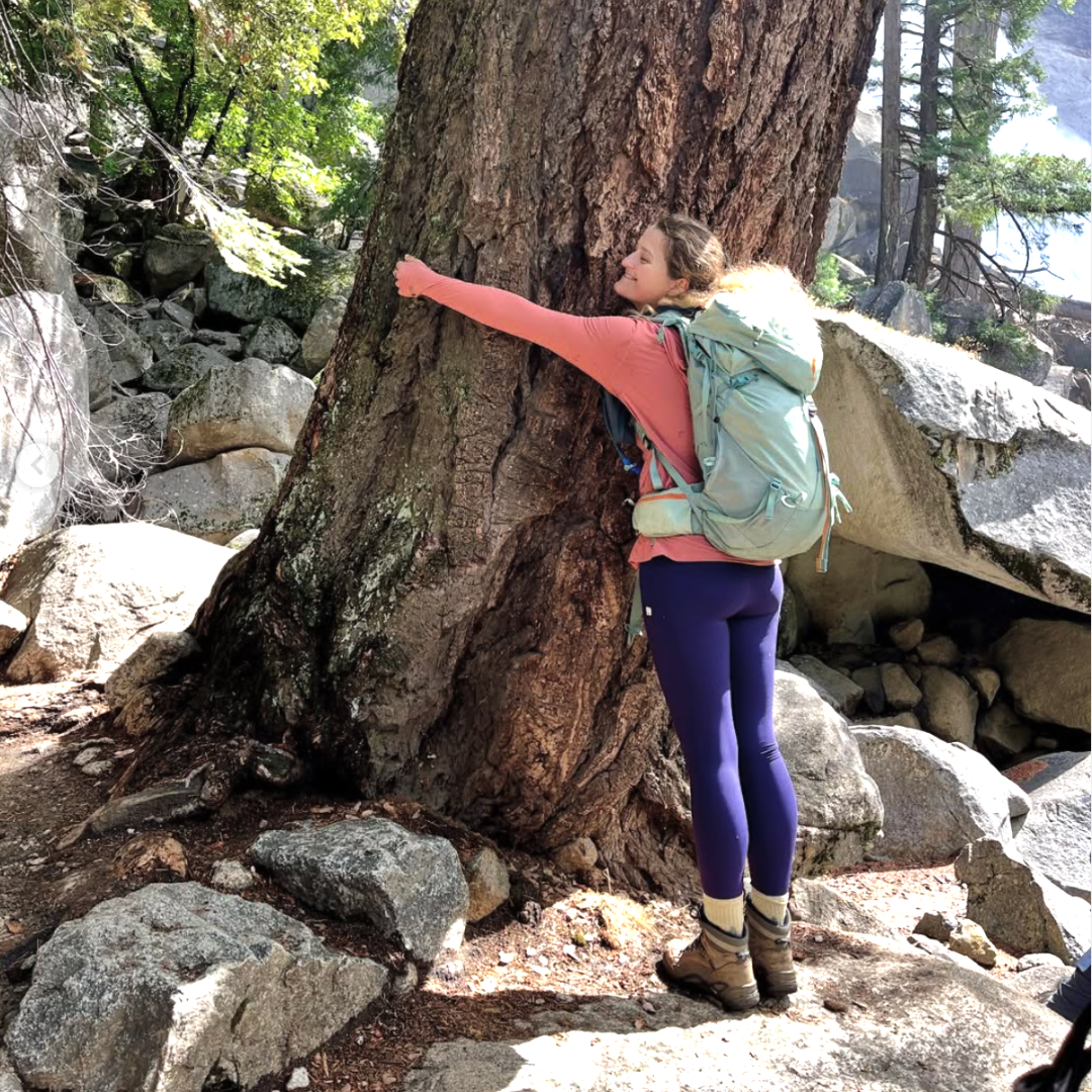 A young woman wears a hiking pack and hugs a tree. Image posted by Instagram user @kdklos. 