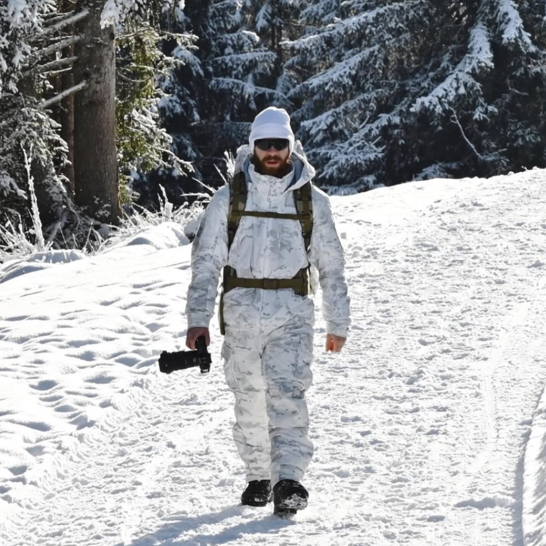 A person in a white camo snow suit walking through the snow holding a camera