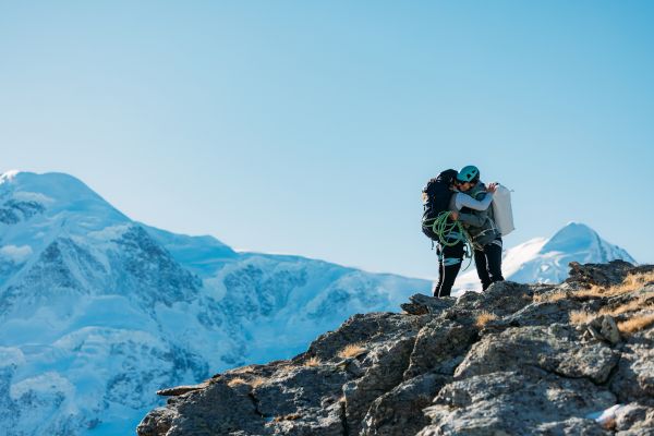 Two women hikers embracing on a mountain peak with snow-capped mountains in the background.